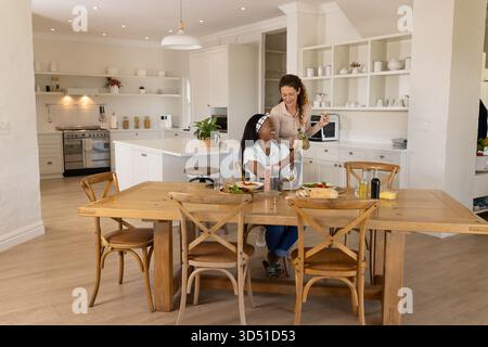 Chatting Diverse female friends sharing salad at wooden dining table in modern kitchen, with wine Stock Photo
