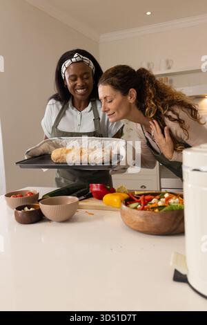 Smelling Diverse female friends leaning on counter in home kitchen, with fresh loaf on baking tray Stock Photo