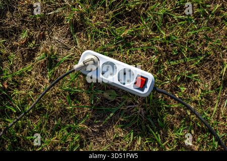 A common white power strip with a red on off switch is sitting on the grass with its black cords going in different directions, selective focus Stock Photo