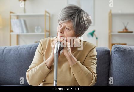 Pensive elderly woman sitting on sofa at home with hands on cane and looking away in deep thought. Stock Photo