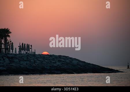 The sun setting directly behind a dark, rocky breakwater, silhouetting palm trees and folded beach umbrellas against a deep orange, hazy sky over the Stock Photo