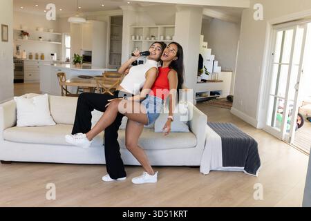 Dancing diverse female friends singing karaoke on light gray sofa in living room, with microphone Stock Photo