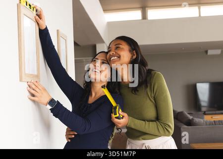 Diverse female friends hanging frame on living room wall using yellow spirit level and tape measure Stock Photo