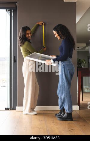 Diverse female friends measuring wall at home, with yellow tape measure and white frame picture Stock Photo