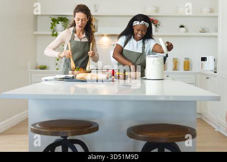 Cooking diverse female friends tossing salad and scooping rice at home kitchen island, rice cooker Stock Photo