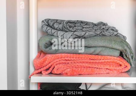A stack of blankets folded on a modern white shelf, including a bright orange plush throw and gray blankets. Stock Photo