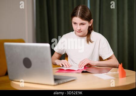 Girl makes origami from colored paper during online video about art of origami, sitting at table and looking at laptop. Online workshop, homework for Stock Photo