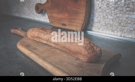 Long wholegrain dark baguette on a wooden cutting board, simple rustic food composition for bakery or culinary themes Stock Photo