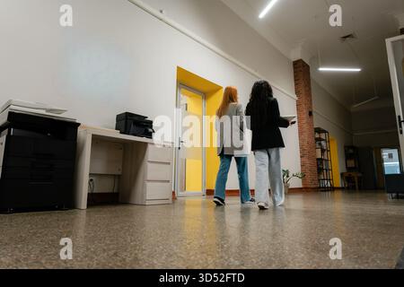 Two colleagues stand in a modern office, discussing near a yellow doorway and brick column Stock Photo