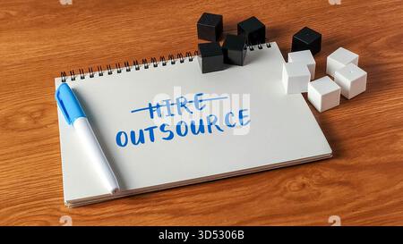 The word Hire crossed out and Outsource written on a notebook with blocks on a wooden desk symbolizing business strategy. Stock Photo