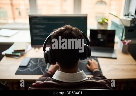 Young adult Caucasian man sitting at desk wearing headphones working on computer with code on monitor, laptop open beside him, focused on programming in modern office setting Stock Photo