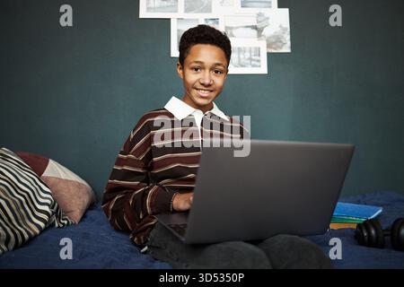 Portrait of Black teenager boy sitting on bed using laptop, smiling at camera, working or studying at home, headphones and books nearby, casual indoor setting, relaxed posture Stock Photo