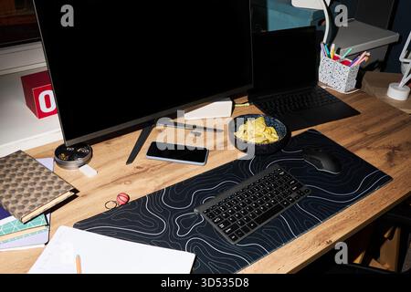 Modern workspace featuring wireless keyboard, computer monitor, laptop, smartphone, bowl of potato chips, stationery items on wooden desk, organized for productivity and remote work Stock Photo