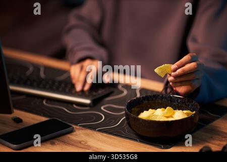 Young adult Black woman working at computer while eating potato chips from bowl, hand holding chip near mouth, smartphone lying on desk, focus on multitasking and snacking Stock Photo