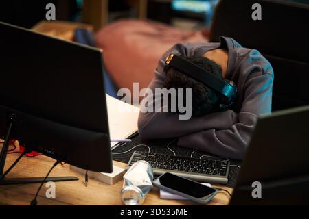Young adult Black man wearing headphones resting head on crossed arms on desk surrounded by computer monitors, keyboard, smartphone and papers, appearing exhausted at workspace Stock Photo