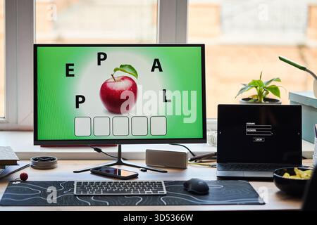 Modern workspace featuring computer monitor displaying word puzzle with apple image and scattered letters, laptop with login screen, smartphone, keyboard, and potted plant on desk Stock Photo