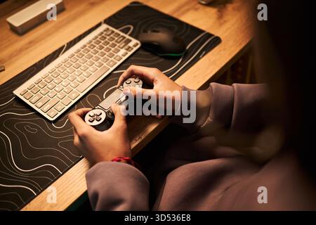 Teenager with medium skin tone holding game controller sitting at desk using computer keyboard and mouse, focusing on gaming activity, hands and partial upper body visible Stock Photo