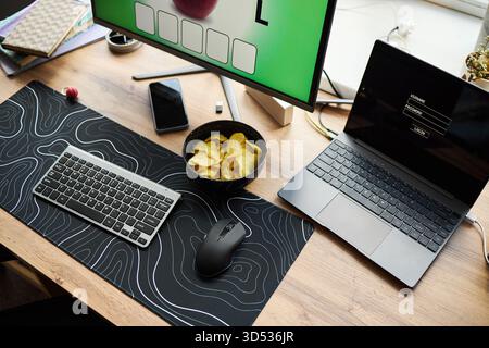 Modern workspace featuring wireless keyboard, computer mouse, laptop with login screen, smartphone, desktop monitor displaying puzzle game, bowl of potato chips on wooden desk Stock Photo