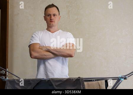 Man standing confidently with arms crossed near loaded drying rack at home. A relaxed man stands with arms crossed Stock Photo