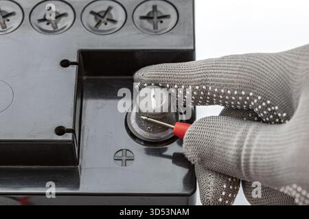 A safety gloved hand uses red multimeter probe to carefully measure the voltage of the positive terminal on a black car battery with visible filler ca Stock Photo