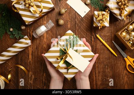 Christmas present packaging. Hands holding a ready-made gift wrapped in white and gold striped paper with a blank tag for your text. Stock Photo