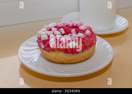 Pink glazed donut with marshmallows on white plate near ceramic coffee cup Stock Photo