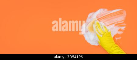 Hand in Yellow Glove Holds Sponge and Wipes Soapy Foam on Orange Background Stock Photo