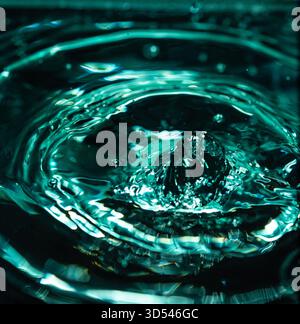 406.A close-up of a water droplet creating a splash and concentric ripples on a dark liquid surface, showcasing the dynamic texture and vibrant teal c Stock Photo