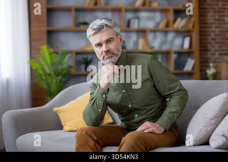 Mature man with stylish grey hair and beard sitting comfortably on sofa in a modern apartment, looking intently at camera with a thoughtful and serious expression on his face Stock Photo