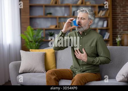 Mature man with white beard clutching his chest on a living room sofa during an asthma attack, using a blue inhaler for urgent breathing relief and medical treatment Stock Photo