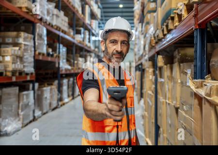 Warehouse worker wearing safety helmet and vest, holding scanner, checking inventory and managing logistics in a large distribution center with packed Stock Photo
