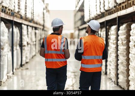 Warehouse workers wearing hard hats and high visibility vests are walking through aisles of stacked inventory, overseeing operations and managing logi Stock Photo
