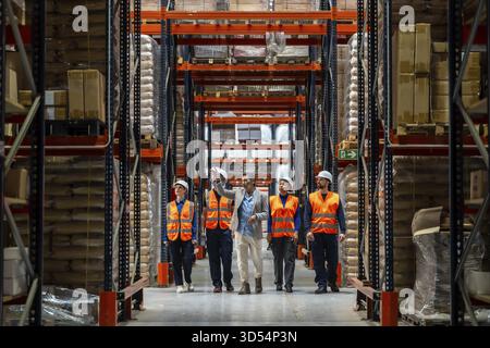Manager discussing inventory and operations with his diverse team of warehouse workers wearing safety vests and hard hats, walking through aisles of g Stock Photo