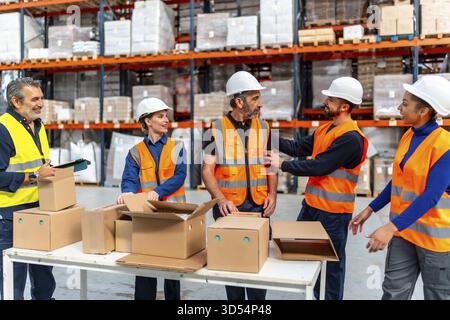 Diverse team of warehouse workers wearing safety vests and hard hats collaborating while packing cardboard boxes on a table in a busy logistics distri Stock Photo