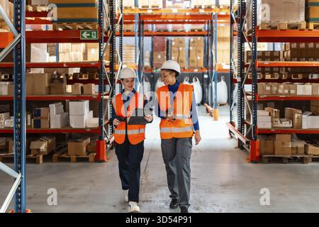 Two diverse female workers wearing safety vests and hard hats, discussing inventory management while walking through a modern logistics warehouse fill Stock Photo