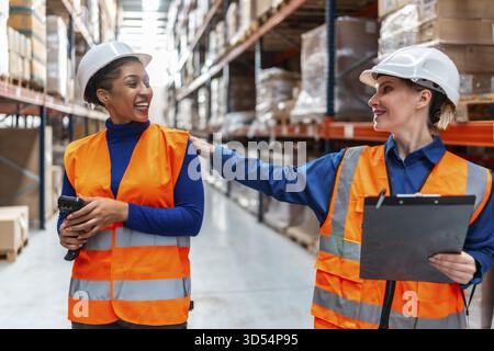 Two diverse women warehouse workers in safety vests and hard hats smiling and collaborating, discussing inventory and operations in a modern logistics Stock Photo