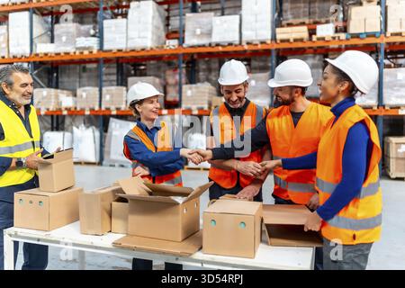 Diverse group of logistics workers shaking hands in a busy distribution warehouse, celebrating a successful collaboration and partnership for efficien Stock Photo