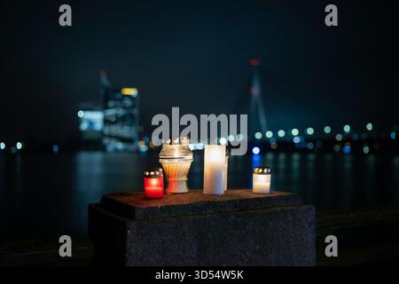 Memorial candles glowing on a stone ledge by the river in Latvia with the illuminated city skyline and distant bridge lights in the background, formin Stock Photo