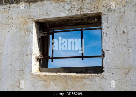 Old wall with window featuring metal bars and a view of clear sky. Textured plaster with cracks framing the rustic scene. Stock Photo