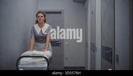 Female Maid in Professional Uniform Moves Cleaning Trolley Down Hallway, Carrying Neatly Stacked Towels For Room Restocking. Concept High Quality Hotel Service, and Efficient Housekeeping Operations. Stock Photo
