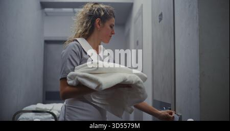 Female Maid in Professional Uniform Moves Cleaning Trolley Down Hallway, Carrying Neatly Stacked Towels For Room Restocking. Concept High Quality Hotel Service, and Efficient Housekeeping Operations. Stock Photo