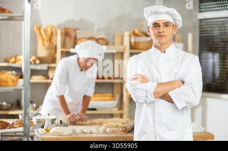 Young baker guy is standing near work table in kitchen, working with dough Stock Photo