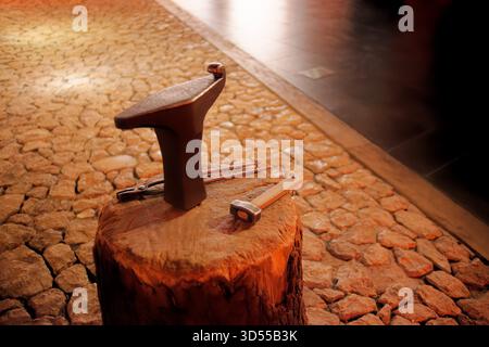 Blacksmith anvil with tools displayed on a wooden block in warm indoor light. Stock Photo