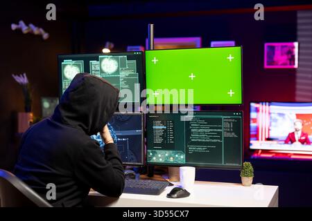 Cybercriminal coding on green screen PC, creating stealth malware that evades security. Hacker using chroma key computer to make script hiding malicious code from detection systems Stock Photo