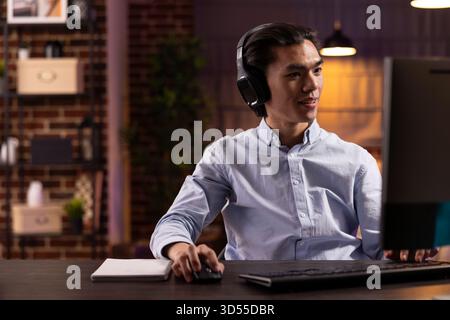 Asian man wearing headphones focusing on computer screen while streaming online webinar. Thoughtful young male adult enjoys learning languages and exploring digital content at home. Stock Photo