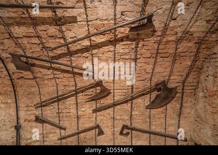 Transylvania, Romania - Oct 8, 2025: Old brick wall with various rusty medieval torture axes and tools hanging on chains, depicting historical Stock Photo