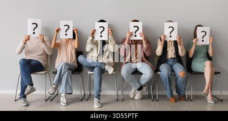 Applicants holding paper sheets with question marks while sitting on chairs near light wall Stock Photo