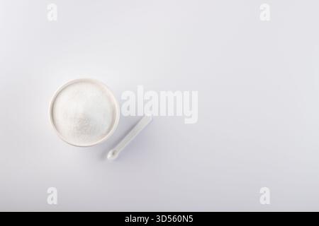 A bowl filled with white sugar sits next to a spoon on a clean, plain surface. This simple setup is perfect for cooking and baking needs in any kitche Stock Photo