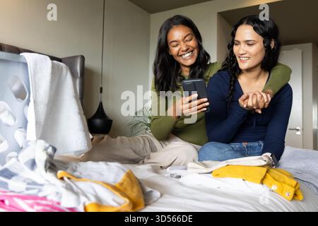 Sitting two women hugging on bed in modern bedroom, with smartphone and laundry basket Stock Photo