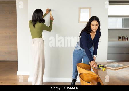 Diverse female friends measuring frames and hammering nails in dining room, with hammer Stock Photo